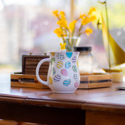 yellow peep bunny hatching from a white egg tea infuser for loose leaf tea with a fine mesh stainless steel tea strainer that reads FLOAT2INFUSE on a green base floating in an easter tea mugh with a vase of flowers in the background
