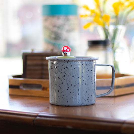red mushroom house tea infuser for loose leaf tea with a fine mesh stainless steel tea strainer that reads FLOAT2INFUSE on a green base floating in a speckled blue tea mug with a vase of flowers in the background