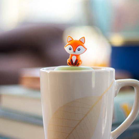 orange fox animal tea infuser for loose leaf tea with a fine mesh stainless steel tea strainer that reads FLOAT2INFUSE on a green base floating in a white tea mug with books in the background