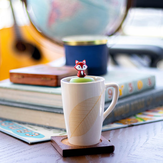 brown fox animal tea infuser for loose leaf tea with a fine mesh stainless steel tea strainer that reads FLOAT2INFUSE on a green base floating in a white tea mug with books in the background