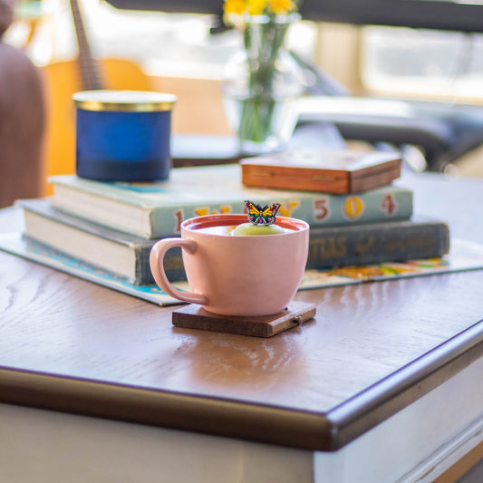 rainbow butterfly animal tea infuser for loose leaf tea with a fine mesh stainless steel tea strainer that reads FLOAT2INFUSE on a green base floating in a pink tea mug with books and a vase of flowers in the background