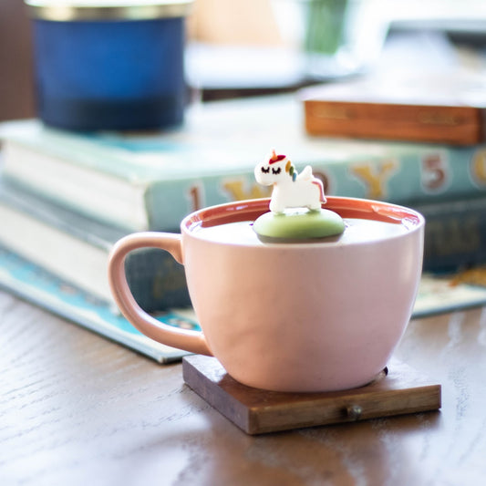 white unicorn with a rainbow mane and gold horn tea infuser for loose leaf tea with a fine mesh stainless steel strainer that reads FLOAT2INFUSE on a green base floating in a pink tea cup with books and a candle in the background