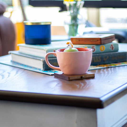 smiling white unicorn with gold mane tea infuser for loose leaf tea with a fine mesh stainless steel tea strainer
that reads FLOAT2INFUSE on a green base floating in a pink tea mug on a table with books, a candle and flowers in the background