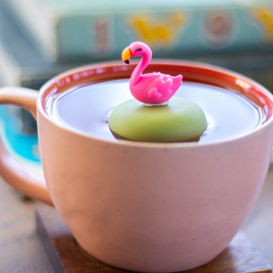 pink flamingo tea infuser for loose leaf tea with a fine mesh stainless steel strainer that reads FLOAT2INFUSE on a green base floating in a pink tea cup with books in the background