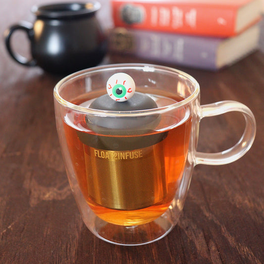 green eyed eyeball tea infuser for loose leaf tea with a fine mesh stainless steel tea infuser that reads FLOAT2INFUSE on a black base floating in a glass tea mug with a black cauldron mug and books in the background