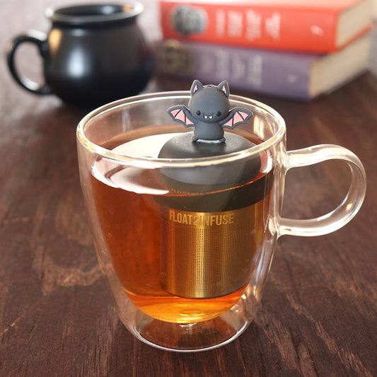 smiling grey and pink bat animal tea infuser for loose leaf tea with a fine mesh stainless steel tea infuser that reads FLOAT2INFUSE on a black base floating in a glass tea mug with a black cauldron mug and books in the background
