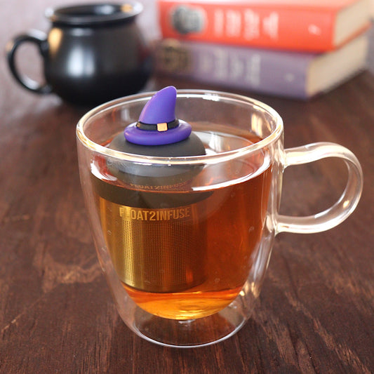 purple wizard hat tea infuser for loose leaf tea with a fine mesh stainless steel tea infuser that reads FLOAT2INFUSE on a black base floating in a glass tea mug with a black cauldron mug and books in the background