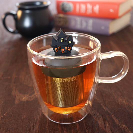 black spooky halloween house tea infuser for loose leaf tea with a fine mesh stainless steel tea infuser that reads FLOAT2INFUSE on a black base floating in a glass tea mug with a black cauldron mug and books in the background