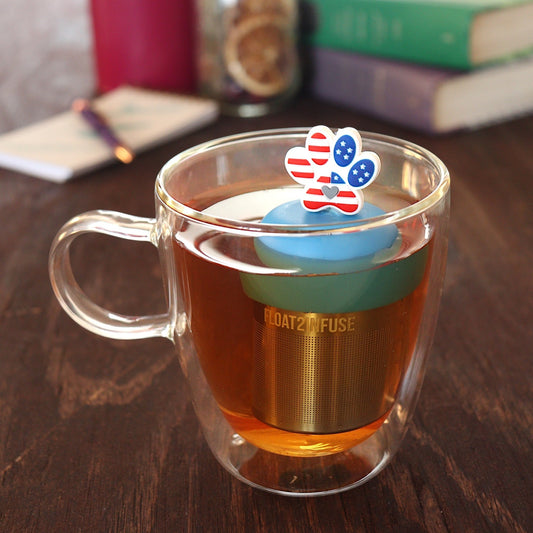 Pawprint with an American flag pattern and grey heart in the center tea infuser for loose leaf tea with a fine mesh stainless steel tea strainer
that reads FLOAT2INFUSE on a blue base floating in a glass tea mug with a pen and notebook, and books in the background