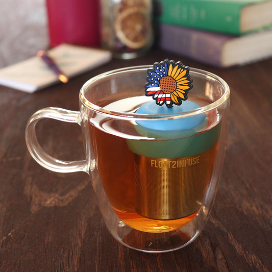 american flag patterned sunflower tea infuser for loose leaf tea with a fine mesh stainless steel tea infuser that reads FLOAT2INFUSE on a blue base floating in a glass tea mug with pen on notebook and loose leaf tea and books in the background