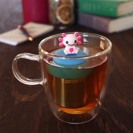 happy pink axolotl tea infuser for loose leaf tea with a fine mesh stainless steel tea strainer that reads FLOAT2INFUSE on a blue base floating in a glass tea mug with a pen on a notebook and books in the background