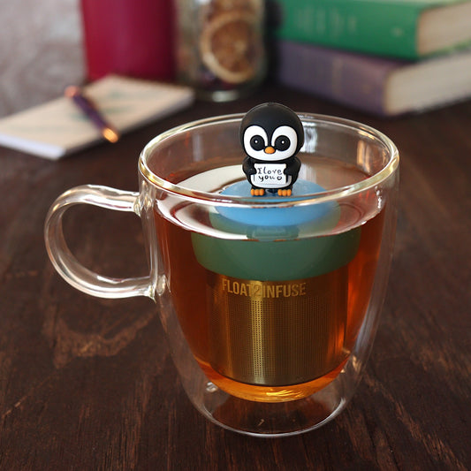 black and white penguin animal holding a sign with text that reads "i love you" tea infuser for loose leaf tea with a fine mesh stainless steel tea infuser that reads FLOAT2INFUSE on a blue base floating in a glass tea mug with pen on notebook and loose leaf tea and books in the background