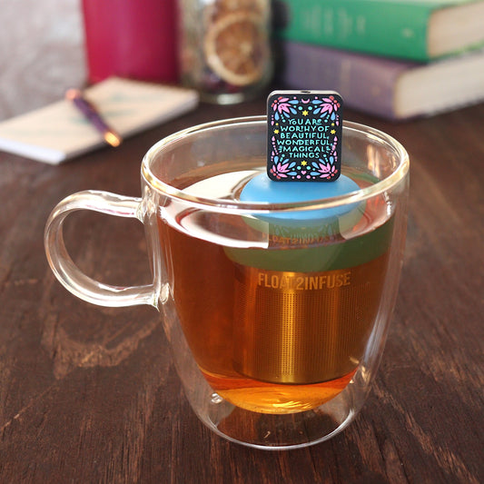 black rectangle with text that reads "you are worthy of beautiful wonderful magical things" tea infuser for loose leaf tea with a fine mesh stainless steel tea infuser that reads FLOAT2INFUSE on a blue base floating in a glass tea mug with pen on notebook and loose leaf tea and books in the background