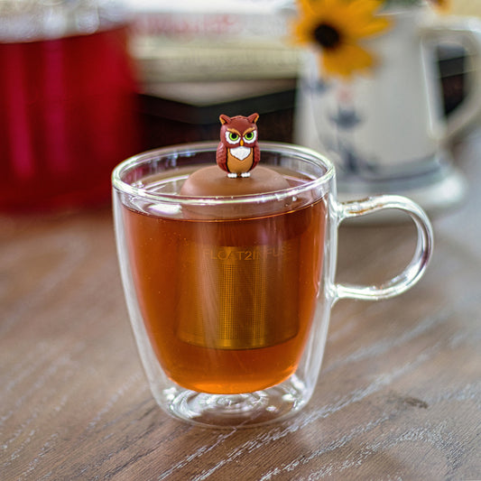 brown owl tea infuser for loose leaf tea with a fine mesh stainless steel tea strainer
that reads FLOAT2INFUSE on a brown base floating in a glass tea mug
with a pitcher and books in the background