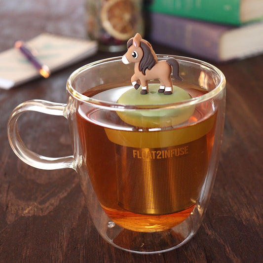 brown horse animal tea infuser for loose leaf tea with a fine mesh stainless steel tea infuser that reads FLOAT2INFUSE on a green base floating in a glass tea mug with pen on notebook and loose leaf tea and books in the background