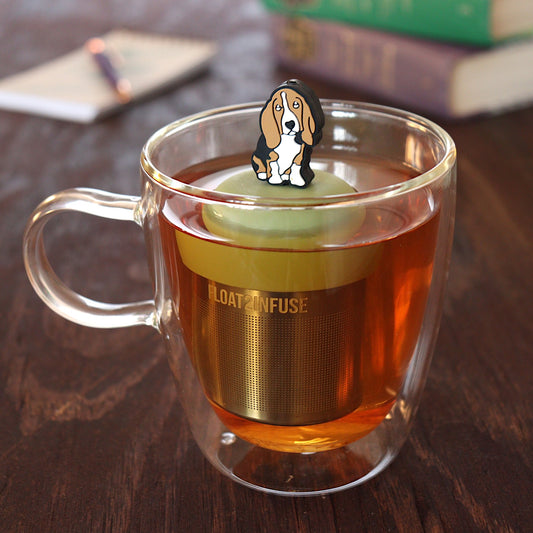 brown and white basset hound dog animal tea infuser for loose leaf tea on a green base with a fine mesh stainless steel tea infuser that reads FLOAT2INFUSE floating in a glass tea mug with pen on notebook and books in the background