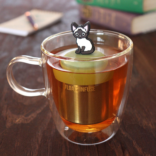 siamese cat animal tea infuser for loose leaf tea on a green base with a fine mesh stainless steel tea infuser that reads FLOAT2INFUSE floating in a glass tea mug with pen on notebook and books in the background