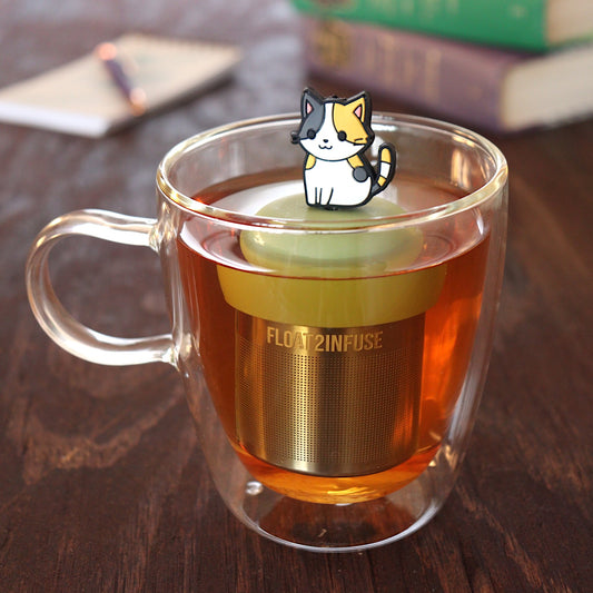 anime Calico cat tea infuser for loose leaf tea on a green base with a fine mesh stainless steel tea infuser that reads FLOAT2INFUSE floating in a glass tea mug with pen on notebook and books in the background