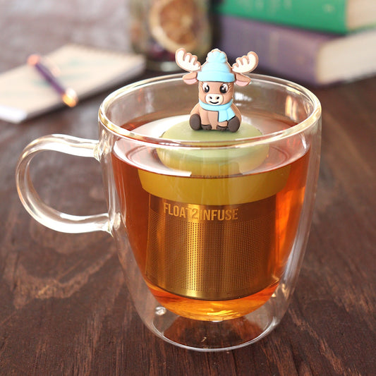 brown reindeer animal wearing blue hat and scarf tea infuser for loose leaf tea with a fine mesh stainless steel tea infuser that reads FLOAT2INFUSE on a green base floating in a glass tea mug with pen on notebook and loose leaf tea and books in the background