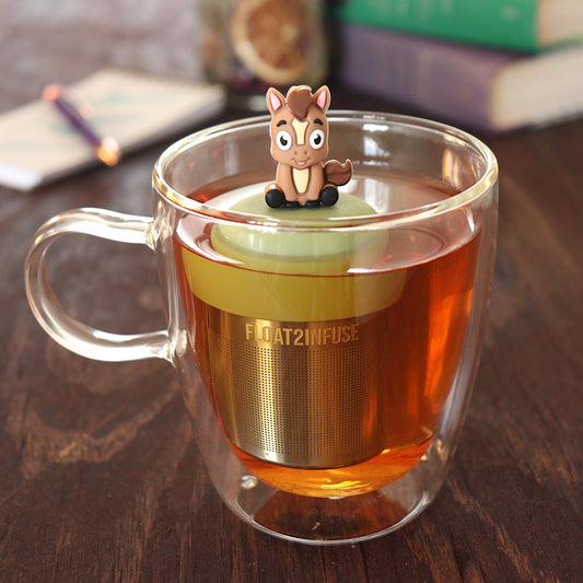 brown donkey animal tea infuser for loose leaf tea on a green base with a fine mesh stainless steel tea infuser that reads FLOAT2INFUSE floating in a glass tea mug with pen on notebook and loose leaf tea and books in the background