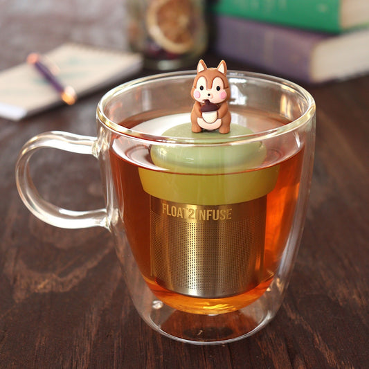 brown chipmunk animal tea infuser for loose leaf tea with a fine mesh stainless steel tea infuser that reads FLOAT2INFUSE on a green base floating in a glass tea mug with pen on notebook and loose leaf tea and books in the background