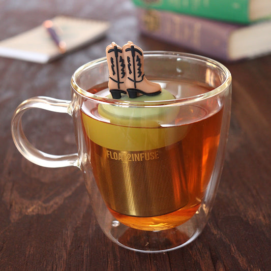 brown cowboy boots tea infuser for loose leaf tea with a fine mesh stainless steel tea infuser that reads FLOAT2INFUSE on a green base floating in a glass tea mug with pen on notebook and books in the background