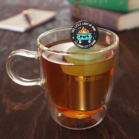 black circle that reads "this little light of mine I'm gonna let it shine" surrounding a smiling dumpster tea infuser for loose leaf tea with a fine mesh stainless steel tea infuser that reads FLOAT2INFUSE on a green base floating in a glass tea mug with pen on notebook and loose leaf tea and books in the background