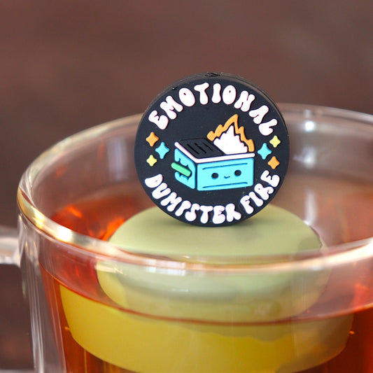 black circle with text that reads "emotional dumpster fire" and an image of a smiling dumpster fire tea infuser for loose leaf tea with a fine mesh stainless steel tea infuser that reads FLOAT2INFUSE on a green base floating in a glass tea mug