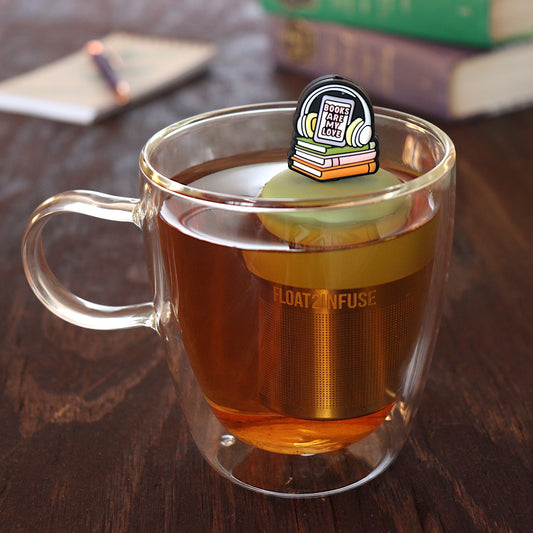 headphones on top of books that reads "books are my love" tea infuser for loose leaf tea with a fine mesh stainless steel tea infuser that reads FLOAT2INFUSE on a green base floating in a glass tea mug with pen on notebook and loose leaf tea and books in the background