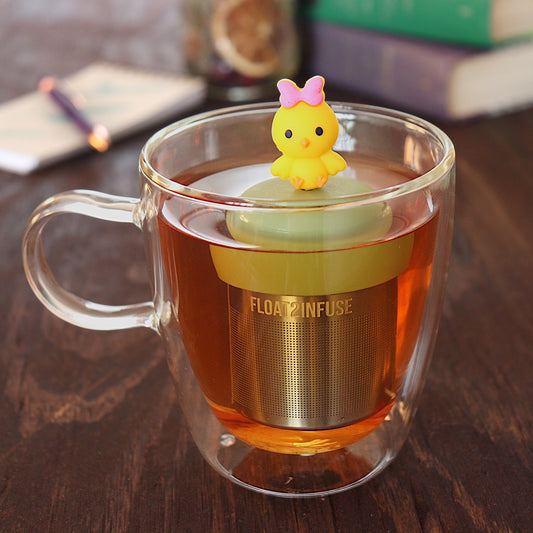 yellow chick with pink bow tea infuser for loose leaf tea with a fine mesh stainless steel tea infuser that reads FLOAT2INFUSE on a green base floating in a glass tea mug with pen on notebook and loose leaf tea and books in the background
