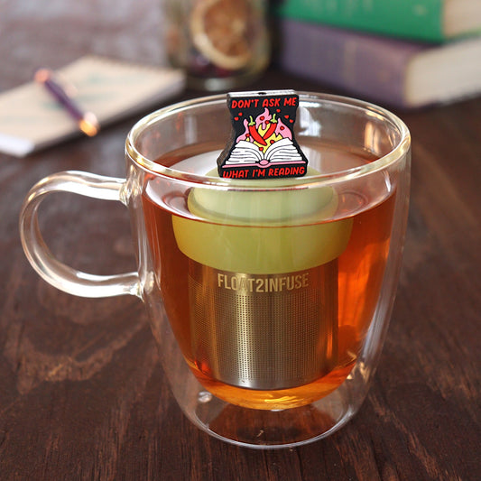 black background with text that reads "don't ask me what I'm reading" with red peppers and flames coming out of an open book tea infuser for loose leaf tea with a fine mesh stainless steel tea infuser that reads FLOAT2INFUSE on a green base floating in a glass tea mug with pen on notebook and loose leaf tea and books in the background