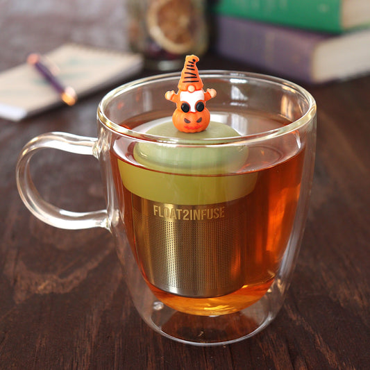 gnome sitting on pumpkin tea infuser for loose leaf tea with a fine mesh stainless steel tea infuser that reads FLOAT2INFUSE on a green base floating in a glass tea mug with pen on notebook and loose leaf tea and books in the background