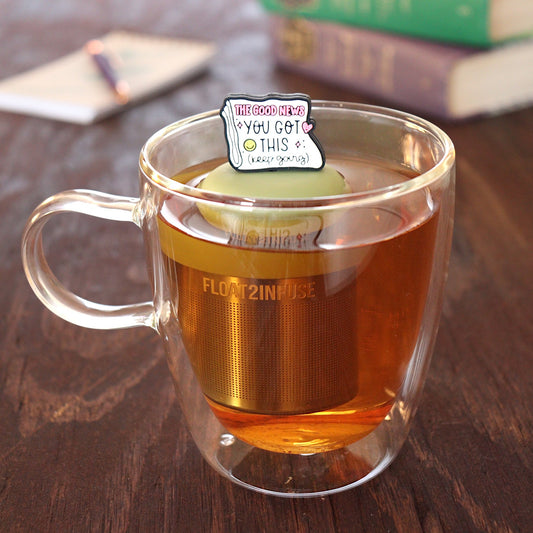 Folded newspaper with text that reads "The good news. You've got this. Keep going" with a pink heart and a yellow smiley face tea infuser for loose leaf tea with a fine mesh stainless steel tea strainer
that reads FLOAT2INFUSE on a green base floating in a glass tea mug with a pen laying on a notebook and books in the background
