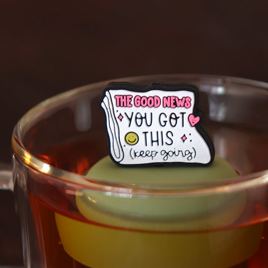 Folded newspaper with text that reads "The good news. You've got this. Keep going" with a pink heart and a yellow smiley face tea infuser for loose leaf tea with a fine mesh stainless steel tea strainer
that reads FLOAT2INFUSE on a green base floating in a glass tea mug