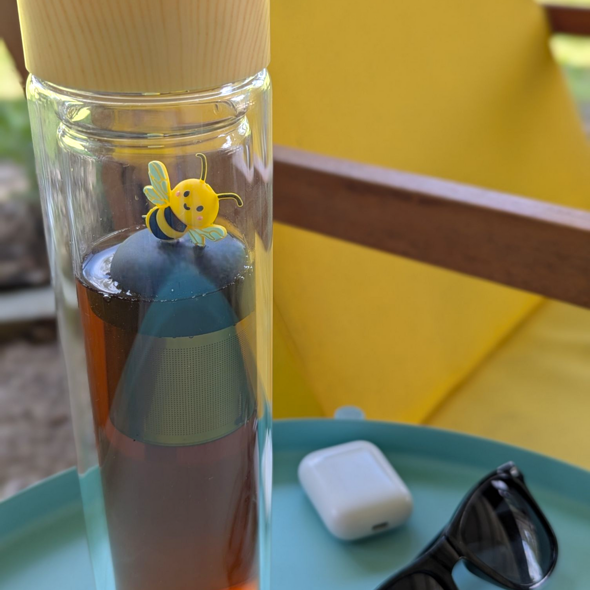 yellow and black bee animal tea infuser for loose leaf tea with a fine mesh stainless steel tea infuser that reads FLOAT2INFUSE on a black base floating in a tall glass travel tea mug with sunglasses, and patio furniture in the background