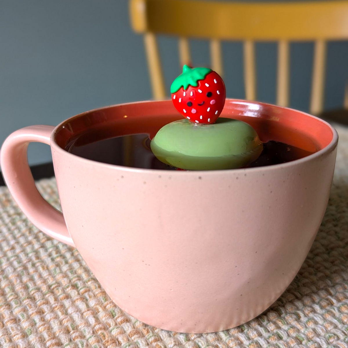 red smiling strawberry tea infuser for loose leaf tea with a fine mesh stainless steel tea strainer that reads FLOAT2INFUSE on a green base floating in a pink tea mug with a chair in the background