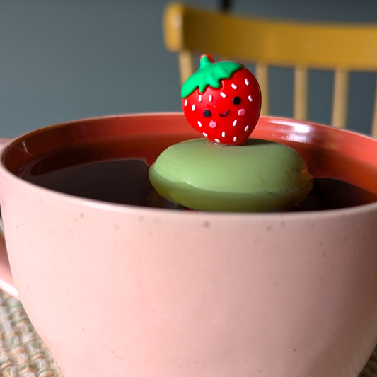 red smiling strawberry tea infuser for loose leaf tea with a fine mesh stainless steel tea strainer that reads FLOAT2INFUSE on a green base floating in a pink tea mug with a chair in the background