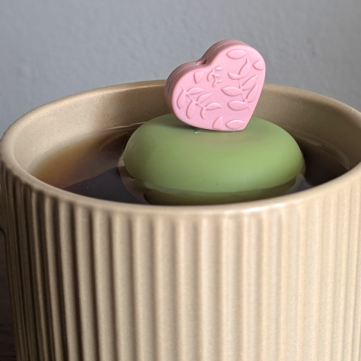 warm pink heart with raised floral pattern tea infuser for loose leaf tea with a fine mesh stainless steel tea strainer that reads FLOAT2INFUSE on a green base floating in a beige tea mug