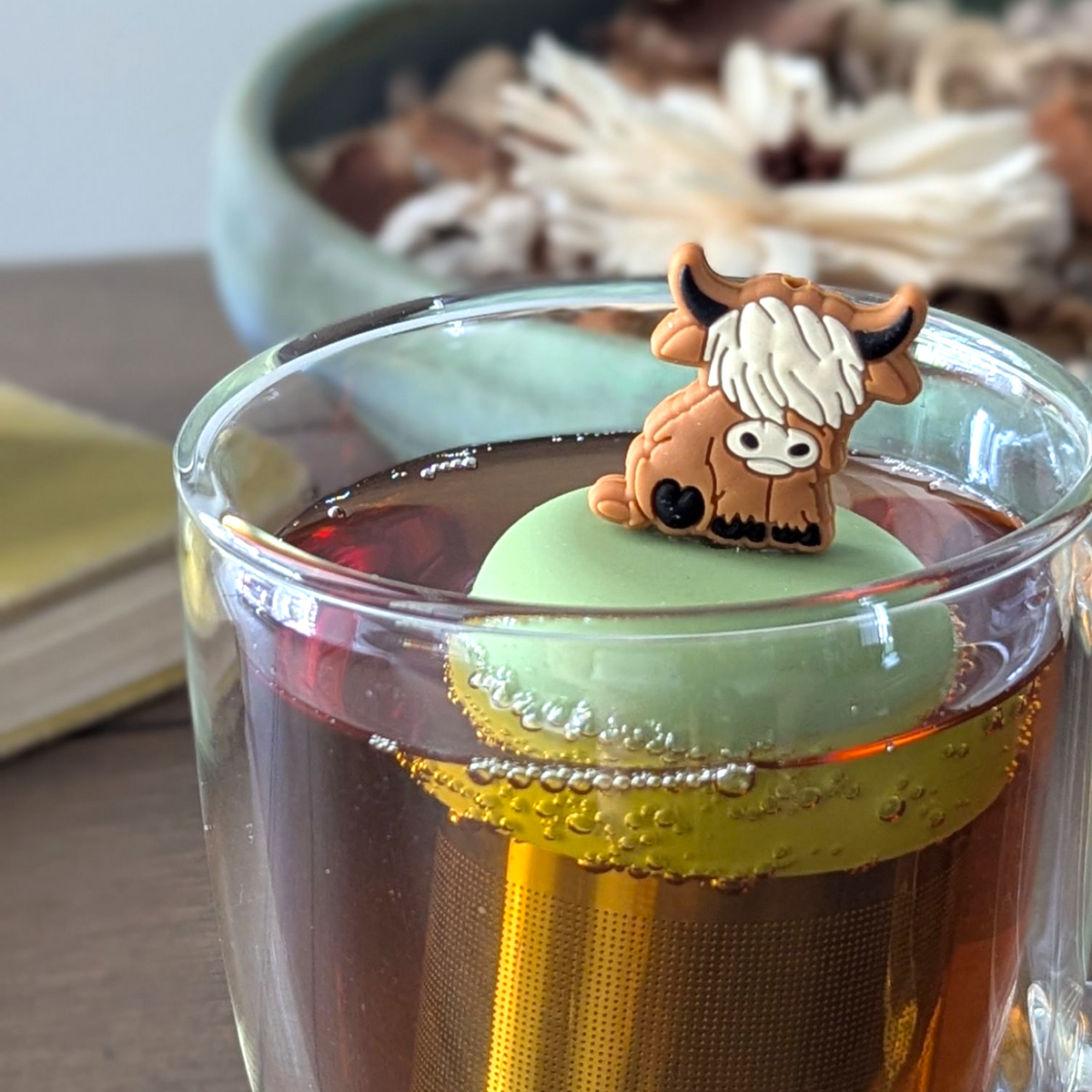 adult brown highland cow animal tea infuser for loose leaf tea with a fine mesh stainless steel tea strainer that reads FLOAT2INFUSE on a green base floating in a glass tea mug with a candle and potpourri in the background