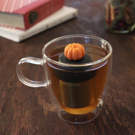 orange pumpkin tea infuser for loose leaf tea with a fine mesh stainless steel tea infuser that reads FLOAT2INFUSE on a black base floating in a glass tea mug with books and loose leaf tea in the background