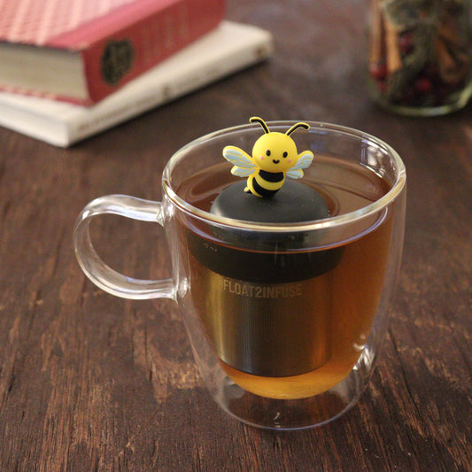 black and yellow bee animal tea infuser for loose leaf tea with a fine mesh stainless steel tea infuser that reads FLOAT2INFUSE on a black base floating in a glass tea mug with books and loose leaf tea in the background
