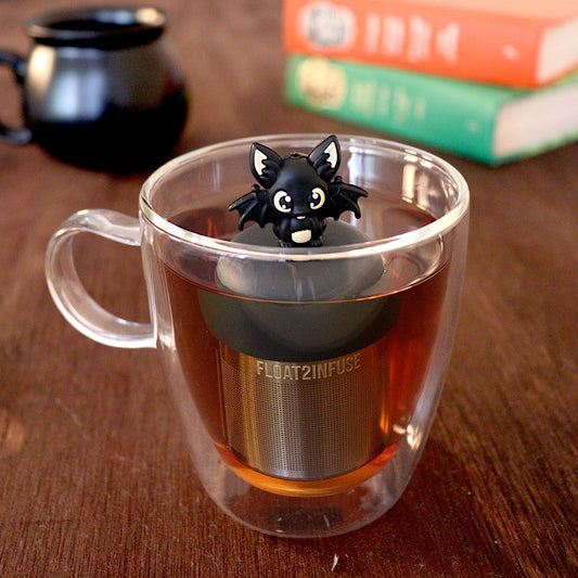 Black bat with outstretched wings tea infuser for loose leaf tea with a fine mesh stainless steel tea strainer
that reads FLOAT2INFUSE on a black base with a black cauldron tea mug and books in the background