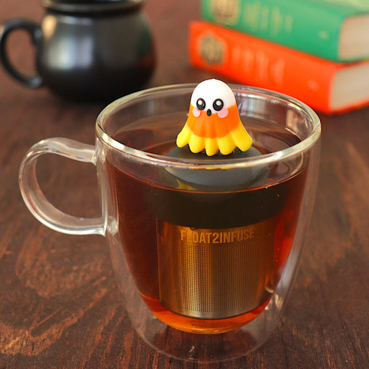 candy corn ghost tea infuser for loose leaf tea with a fine mesh stainless steel tea infuser that reads FLOAT2INFUSE on a black base floating in a glass tea mug with books in the background