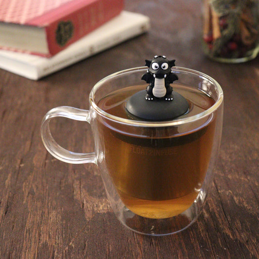 black dragon animal tea infuser for loose leaf tea with a fine mesh stainless steel tea infuser that reads FLOAT2INFUSE on a black base floating in a glass tea mug with books and loose leaf tea in the background