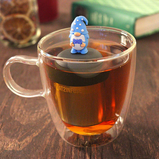 gnome with long white beard wearing blue with white polka dots holds text "dad" tea infuser for loose leaf tea with a fine mesh stainless steel tea infuser that reads FLOAT2INFUSE on a black base floating in a glass tea mug with loose leaf tea and books in the background