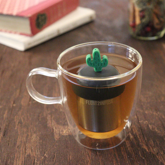 green cactus tea infuser for loose leaf tea with a fine mesh stainless steel tea infuser that reads FLOAT2INFUSE on a black base floating in a glass tea mug with books and loose leaf tea in the background