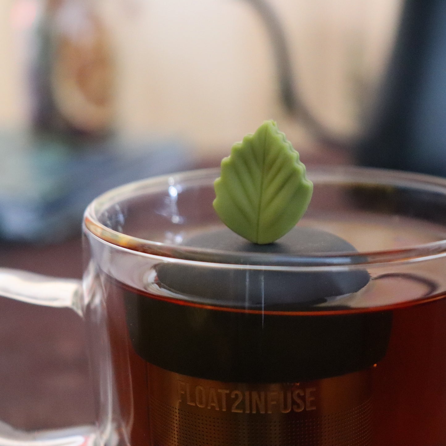 green herb leaf tea infuser for loose leaf tea on a black base with a fine mesh stainless steel strainer that reads FLOAT2INFUSE floating in a glass tea mug with tea kettle in the background