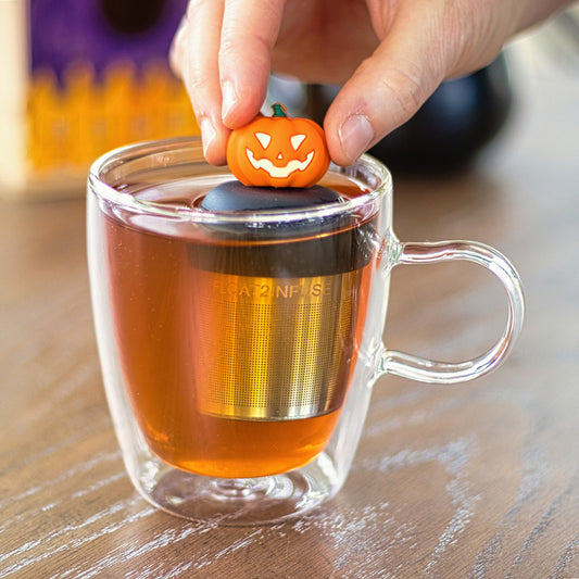 orange jack-o-lantern tea infuser for loose leaf tea with a fine mesh stainless steel tea strainer that reads FLOAT2INFUSE on a black base floating in a glass tea mug with a cauldron and books in the background