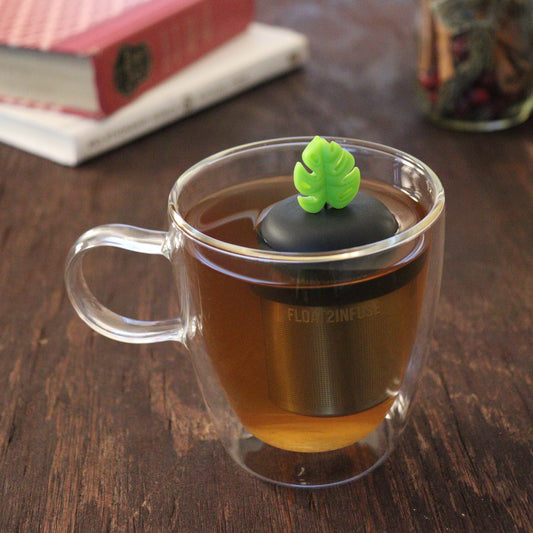 lime green monstera leaf tea infuser for loose leaf tea with a fine mesh stainless steel tea infuser that reads FLOAT2INFUSE on a black base floating in a glass tea mug with books and loose leaf tea in the background