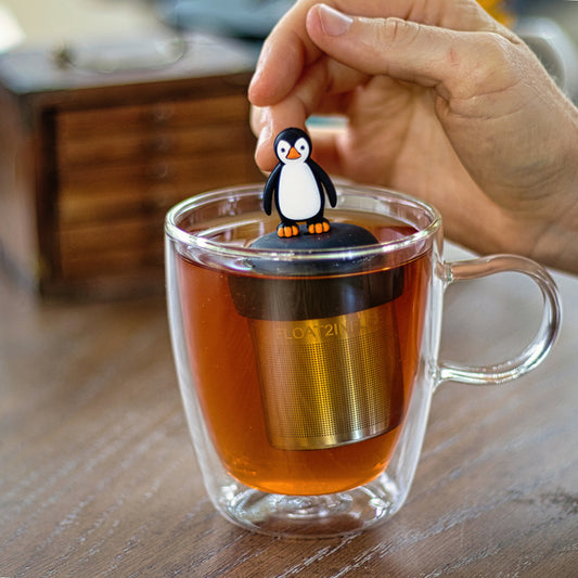 black and white penguin tea infuser for loose leaf tea with a fine mesh stainless steel strainer that reads FLOAT2INFUSE on a black base floating in a glass tea mug with wooden box in the background